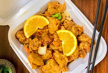 Crispy Orange Chicken in a takeout container, with orange slices, veggies, and chopsticks on a wooden surface at Chopsticks，a Chinese Restaurant in Kent