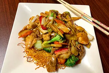 A plate of Hong Kong Noodle with stir - fried chicken, assorted vegetables like broccoli, bell peppers, and mushrooms, served over crispy noodles. A pair of decorated chopsticks is placed beside it on a wooden table at Chopsticks，a Chinese Restaurant in Kent