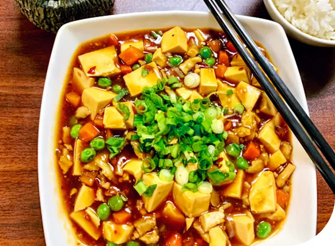 A plate of Mapo Tofu, with soft tofu cubes in a rich, savory sauce, mixed with peas, diced carrots, and minced meat. Topped with chopped green onions, served with chopsticks, and accompanied by a bowl of white rice on a wooden table at Chopsticks，a Chinese Restaurant in Kent