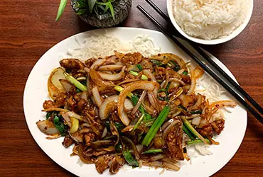 A plate of Mongolia Beef with tender beef strips, stir - fried onions and green onions, served over noodles. Accompanied by a bowl of white rice and chopsticks on a wooden table, with a small potted plant in the background at Chopsticks，a Chinese Restaurant in Kent