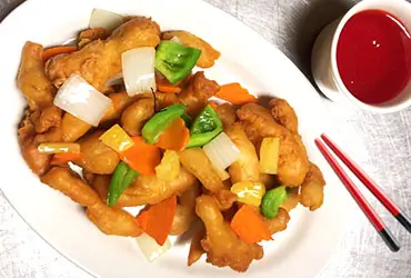 A plate of Sweet Sour Chicken with crispy chicken pieces, mixed with colorful bell peppers, onions, and pineapple chunks. Served with a small bowl of red dipping sauce and a pair of red chopsticks on a light - colored surface at Chopsticks，a Chinese Restaurant in Kent