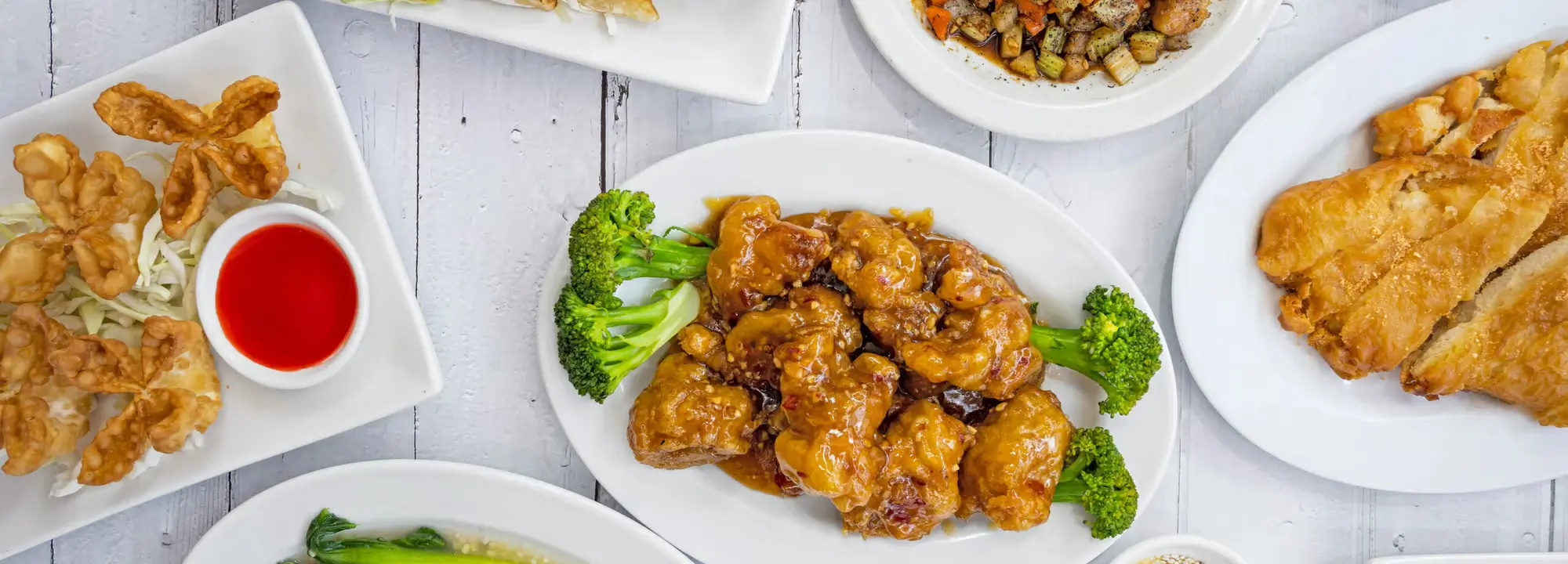 A plate of glazed chicken pieces, surrounded by broccoli florets, presented on a white plate among other dishes on a light - colored wooden table at Chopsticks，a Chinese Restaurant in Kent